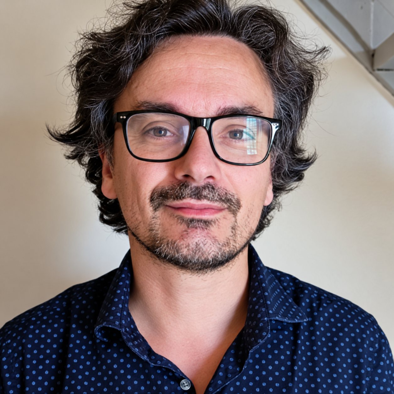 Portrait d’un homme aux cheveux bruns bouclés, avec des lunettes noires et une chemise bleu foncé